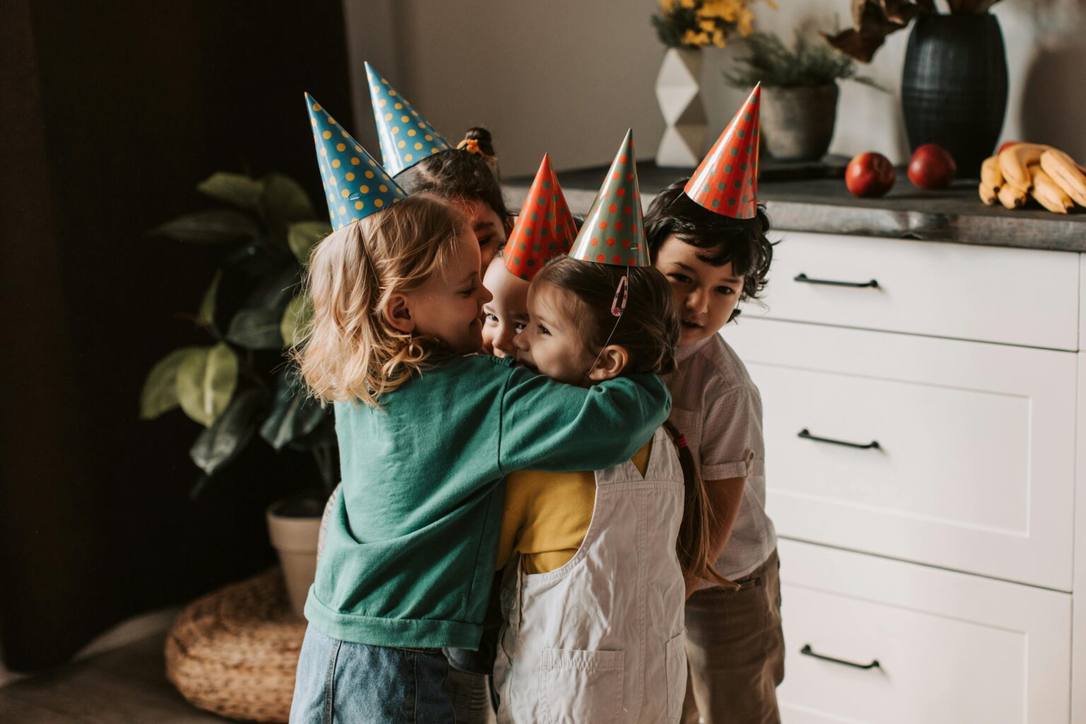 Four kids wearing party hats and hugging warmly during a birthday celebration indoors.