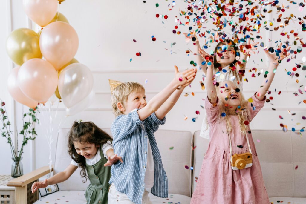 A group of children celebrating a birthday party with colorful balloons and confetti indoors.
